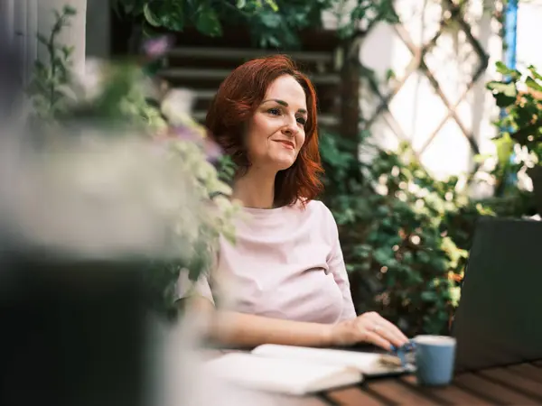 Smiling woman interacting with laptop on a plant-decorated balcony, exuding a sense of calm and contentment