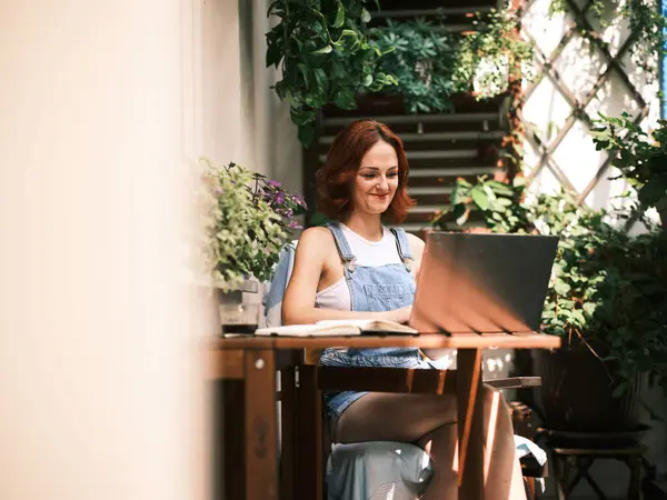 Leende Kvinna Jeansdräkt Arbetar Sin Laptop Omgiven Växter Solig Balkong Stockbild