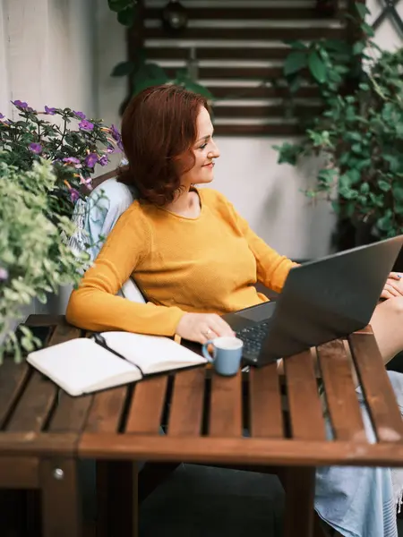 一位红头发的年轻女子喜欢在阳光灿烂的阳台上 在她的笔记本电脑上工作 身边围着茂盛的盆栽 — 图库照片