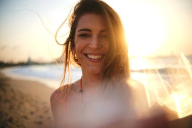 Gün batımında kumsalda kameraya gülümseyen genç bir kadının güneşli portresi.