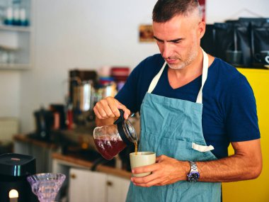 Deniz mavisi önlük giyen bir barista kahve tezgahındaki beyaz bardağa taze kahve döker. Bu da davetkar bir atmosfer ve bir kafede kaliteli bir hizmete adanmışlığı tasvir eder..