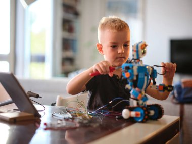 Genç bir çocuk tamamen renkli bir robota kablolar bağlamaya odaklanmış, masadaki aletlerle bu aktif aktiviteyle uğraşırken yaratıcılık ve konsantrasyonu göstermeye çalışıyor..