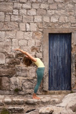 Sarı bluz ve yeşil tayt giymiş bir kadın deniz kenarında savaşçı yoga pozu veriyor. Görkemli dağlar ve arka planda açık gökyüzü var. Gücü arttırıyor..