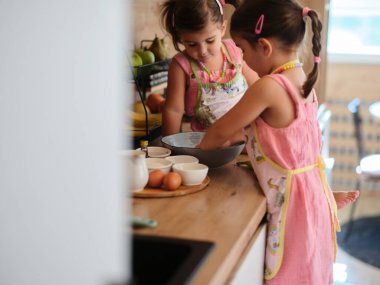 Mutfakta, önlüklü iki hevesli kız birlikte yemek hazırlarken, çeşitli yemek malzemeleriyle çevrelenmiş, takım çalışması ve öğrenme gösterileri yaparken görülüyor..