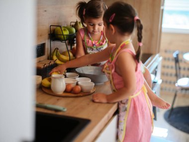 Önlük takan iki genç kız, muzlar, yumurtalar ve diğer malzemelerle çevrili bir mutfakta birlikte yemek pişiriyor ve eğlenceli bir işbirliği anı yakalıyorlar..