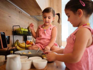 Önlüklü iki kız kardeş, annelerinin dikkati altında muzlu, yumurtalı ve çeşitli malzemelerle yemek hazırlamaktan zevk alırlar. Eğlenceli ve eğitici yemek pişirme aktiviteleri sergilerler..