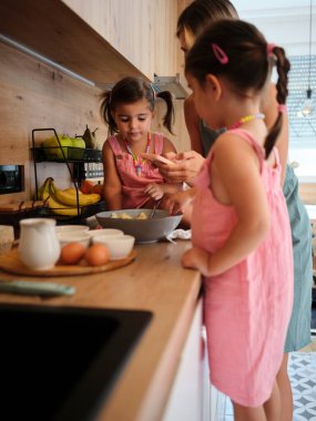 Önlüklü iki küçük kız, annelerinden yardım alarak muz, yumurta ve diğer önemli şeylerle dolu sıcak bir mutfakta yemek hazırlıyorlar. Takım çalışmasını ve eğlenceyi sembolize ediyorlar..