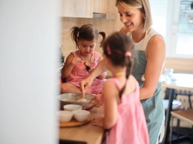 Bir anne, sıcak mutfakta kızlarına yemek pişirmede yardım ediyor. Sıcak ve sevgi dolu bir ortam yaratıyor. Birlikte yemek tarifi yapmaktan hoşlanıyorlar..