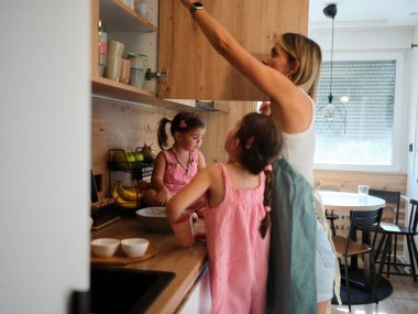 Bir aile pişirme seansı, bir anne ve iki kızının modern bir mutfakta yemek pişirmesi, aile birliğini ve öğretim anlarını göstermesi ile resmedilir..