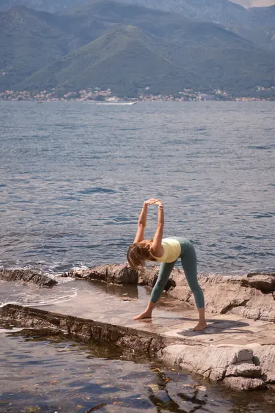 stock image A woman in yellow top and green leggings holds a plank pose on a rocky shoreline with mountains and cloudy sky in the background, showing strength and determination.