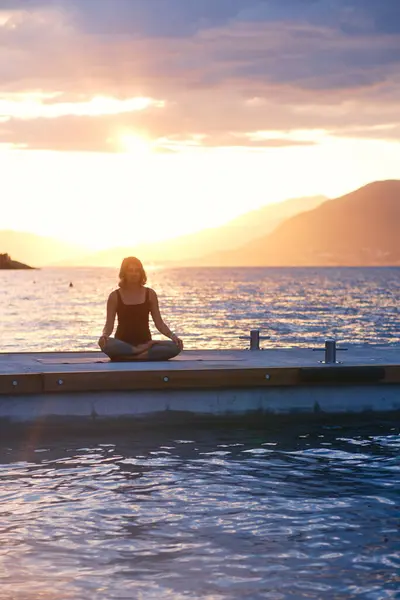 stock image A woman in yellow top and green leggings holds a plank pose on a rocky shoreline with mountains and cloudy sky in the background, showing strength and determination.