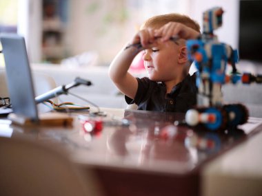 Bir çocuk, yakınlarda dizüstü bilgisayarı olan bir robotu bir araya getirmekle derinden meşguldür, modern bir evde odaklanmış bir merak ve elle öğrenme anını yakalar..