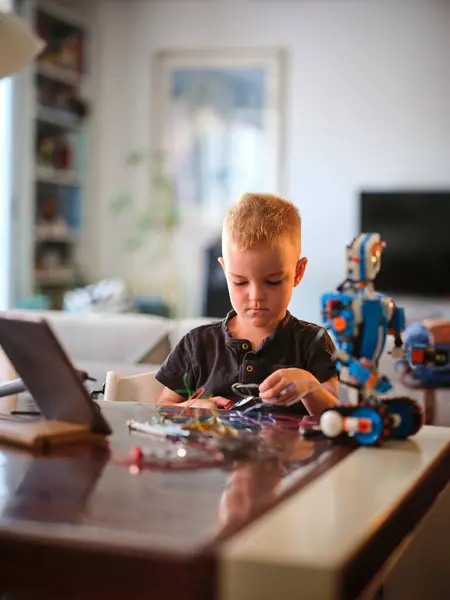 stock image A young boy's furrowed brow reflects serious concentration as he wires a blue robot, surrounded by colorful wires on a table in a vibrant home environment.