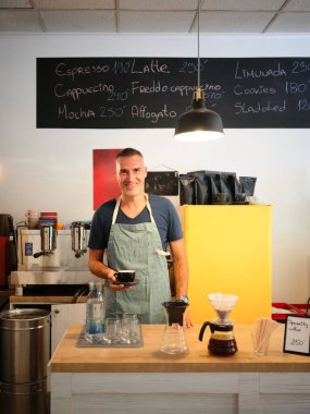 Yeşil önlüklü bir erkek barista kahve tezgahının arkasında elinde bir fincan kahveyle duruyor. Çekici ve düzenli bir şekilde hazırlanan kafede servis yapmaya hazır..