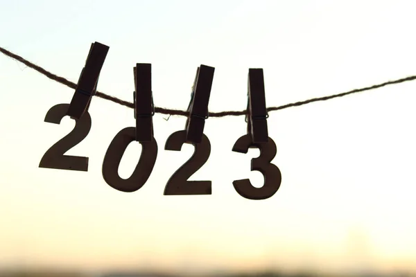 木製の番号は オレンジ色の朝の空の日の出とぼやけた都市のボケ光の背景の上に服のペグピンで接続されたロープにハングアップします 明るいスタート日のコンセプトのための幸せな新年2023お祝い — ストック写真