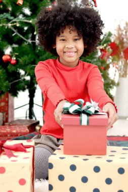 Siyah köri saçlı, gülümseyen Afro-Amerikan kızın portresi Noel hediyesi kutusunu dekoratif Noel ağacının altında tutuyor, çocuk Noel 'in mutlu kış tatilini kutluyor