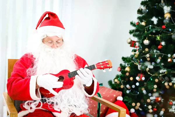 Santa Claus sitting on chair and playing Ukulele, singing Christmas songs front of decorative Christmas tree in living room, Merry Christmas happy winter holiday celebration.