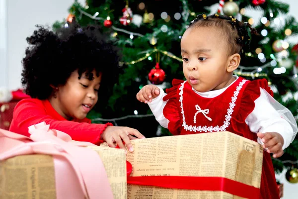 Two adorable happy smiling African American little girl sisters with black curry hair hold many gift boxes presents under Christmas tree in living room, kid celebrating happy Christmas winter holiday