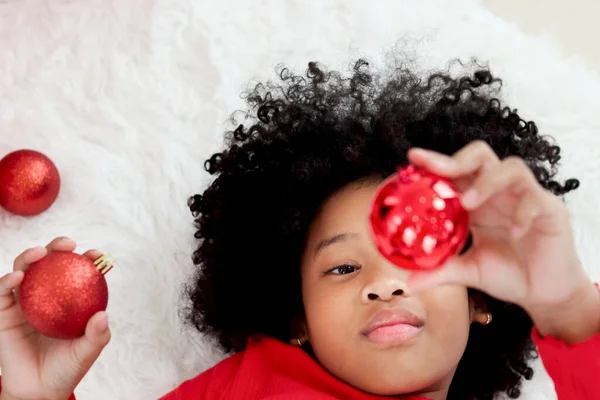 Top view adorable happy African American girl child with black curry hair lying down on white carpet at living room floor, holding Christmas ball ornament and reaching it up, Christmas winter holiday