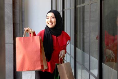 Mutlu gülümseyen Asyalı genç Müslüman kadınlar kırmızı başörtüsü takmış ellerinde alışveriş çantalarıyla caddede yürüyen güzel bir kızın portresi.