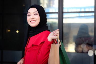 Mutlu gülümseyen Asyalı genç Müslüman kadınlar kırmızı başörtüsü takmış ellerinde alışveriş çantalarıyla caddede yürüyen güzel bir kızın portresi.