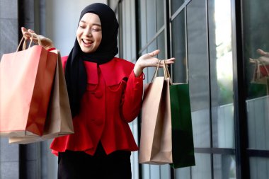 Mutlu gülümseyen Asyalı genç Müslüman kadınlar kırmızı başörtüsü takmış ellerinde alışveriş çantalarıyla caddede yürüyen güzel bir kızın portresi.