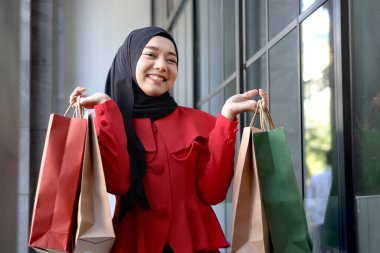 Mutlu gülümseyen Asyalı genç Müslüman kadınlar kırmızı başörtüsü takmış ellerinde alışveriş çantalarıyla caddede yürüyen güzel bir kızın portresi.