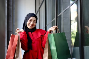 Mutlu gülümseyen Asyalı genç Müslüman kadınlar kırmızı başörtüsü takmış ellerinde alışveriş çantalarıyla caddede yürüyen güzel bir kızın portresi.