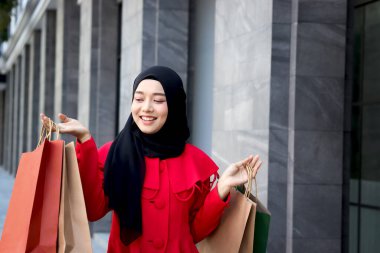 Mutlu gülümseyen Asyalı genç Müslüman kadınlar kırmızı başörtüsü takmış ellerinde alışveriş çantalarıyla caddede yürüyen güzel bir kızın portresi.