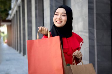Mutlu gülümseyen Asyalı genç Müslüman kadınlar kırmızı başörtüsü takmış ellerinde alışveriş çantalarıyla caddede yürüyen güzel bir kızın portresi.