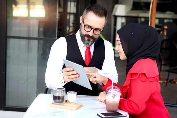 Senior elderly mature business man using tablet and discussing with hijab Muslim women during having a coffee at outdoor cafe, happy harmony people take a break from drinking beverage, enjoy relaxing.