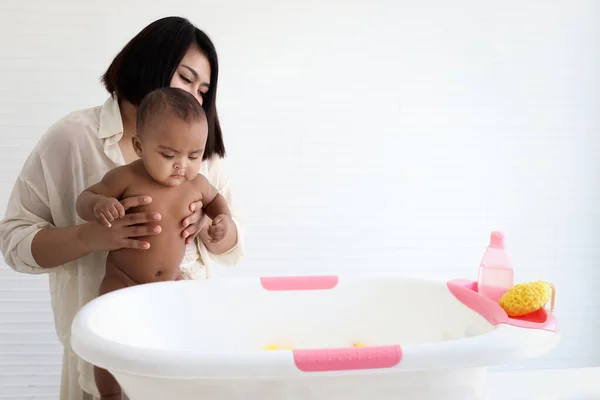 Mother holding six month Asian African American infant baby to take bubble bath in bathroom, mother bathing kid in tub, mom washing and cleaning her child in bath.