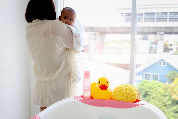 Yellow duck toy, shampoo soap and sponge (baby toiletries) on bathtub in bathroom with blurry background of mother holding infant baby covered with white towel.