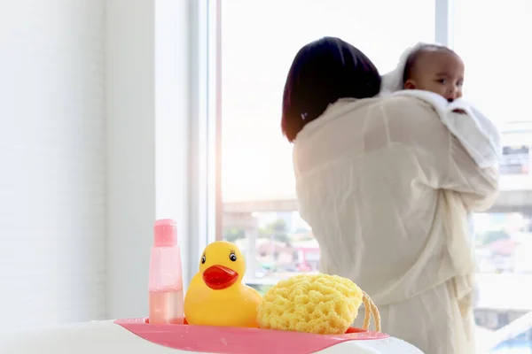 Yellow duck toy, shampoo soap and sponge (baby toiletries) on bathtub in bathroom with blurry background of mother holding infant baby covered with white towel.