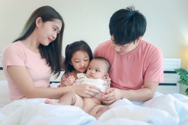 Mutlu Asyalı aile yatak odasında eğleniyor. Baba, anne, kız ve bebek yatakta birlikte yatıyorlar, abla küçük bebeği tutmaya çalışıyor. Anne ve baba, ailelerin çocukla arası iyidir.