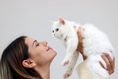 Genç, mutlu, gülümseyen, sevimli, beyaz bir İran kedisi tutan, kedisini kaldıran, evcil hayvanını öpmeye çalışan, tüylü, uzun saçlı, sevimli kedisine sevgiyle sarılan bir kız.