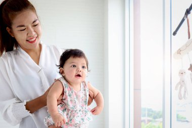 Happy smiling adorable six months baby girl standing next to the window in embrace of mother arms, mom holding her sweet little daughter kid.