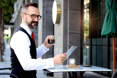 Üst düzey olgun iş adamı dışarıda kahve içerken dijital masasını kullanıyor, mutlu insanlar içecek içmeye ara veriyor, rahatlamanın tadını çıkarıyor..