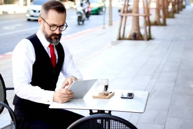 Senior elderly mature business man using digital table stressful thinking about work during having coffee at outdoor cafe, serious man overthinking during taking a break from drinking beverage.