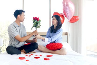 Happy Asian couple hold red rose bouquet, look at each other while sit on white bed in bedroom, romantic man give beautiful flower to girlfriend, lover celebrate memorable anniversary on Valentine day