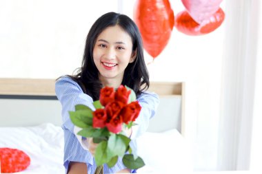 Beautiful red rose bouquet on Asian woman hand, happy girl showing Valentine flower present from lover boyfriend to camera while sitting on white bed in bedroom. Happy Saint Valentine Day.