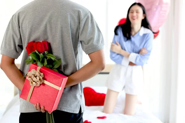 Man hiding red gift box with beautiful red rose bouquet behind back, ready to give Valentine present to lover girlfriend for celebration, couple make surprise, love and happiness. Happy Valentine Day