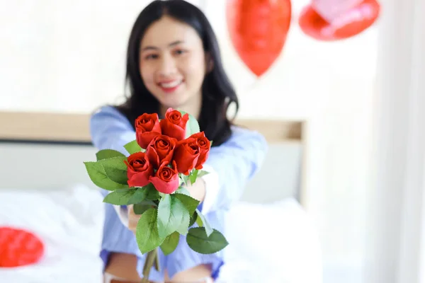 Beautiful red rose bouquet on Asian woman hand, happy girl showing Valentine flower present from lover boyfriend to camera while sitting on white bed in bedroom. Happy Saint Valentine Day.