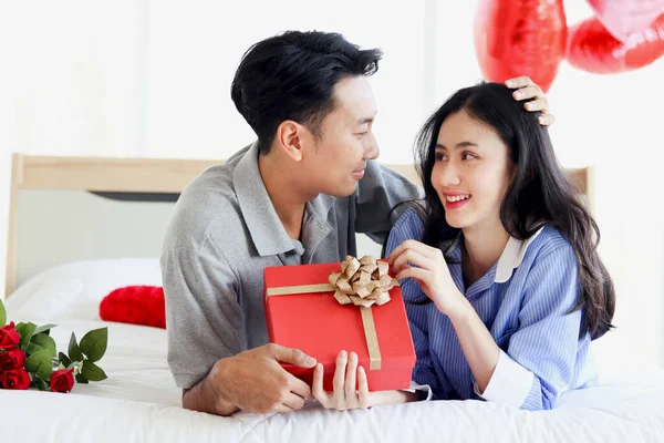 Happy Asian couple looking at each other while lying on white bed in bedroom, romantic man giving red Valentine gift box present to his girlfriend to celebrate memorable anniversary on Valentine Day
