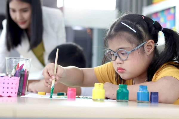 Happy children have fun with friends during study at school, girl with down syndrome concentrate painting on paper in art classroom, education of kids with physical disability and intellectual concept