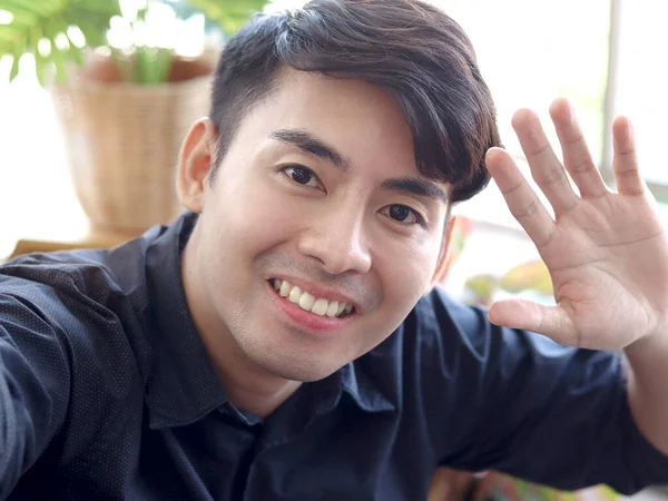 Young handsome Asian man in black shirt waving hand in greeting gesture by using smartphone to social media meeting online, communicating on video call, use mobile phone for selfie in living room.