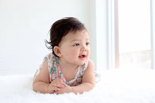 Portrait of six months crawling lovely baby on fluffy white rug next to the window with copy space, happy smiling sweet little cute girl kid lying on bed in bedroom, childhood and baby care concept
