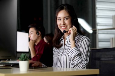 Mutlu gülümseyen, kulaklıklı güzel Asyalı bir kadının portresi çağrı merkezi müşteri hizmetleri masasında gece vardiyasında çalışıyor, kameraya bakıyor, cep telefonsuz çalışan çok çalışan bir kadın..