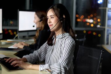 Kulaklıklı güzel Asyalı kadın gece vardiyasında çalışıyor. Müşteri hizmetleri masasında. Masaüstü bilgisayarına bakıyor..