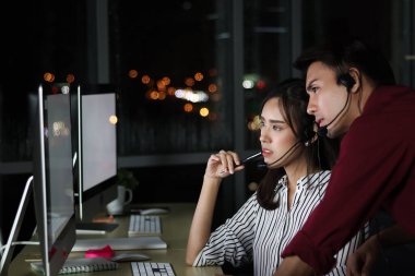 Kulaklıklı Asyalı gece vardiyası çalışanları tartışma için birlikte masaüstü bilgisayarına bakarlar. Gece vardiyasında çalışan, elleri boş çalışan ve müşteri hizmetlerinde fazla mesai yapan bir ekip.